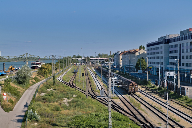 Ein Zug fährt auf Schienen neben einer Stadtlandschaft mit Strommasten, Gebäuden, Grün, Verkehrsschildern, Fahrzeugen, einer Brücke, Wasser und einem bewölktem Himmel.