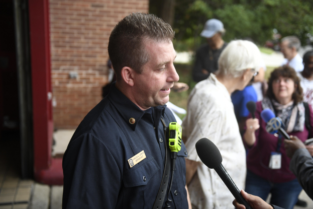 Ein Polizist in Uniform, der mit einer Gruppe von Menschen spricht, mit Mikrofonen, Bäumen und einem Gebäude im Hintergrund.