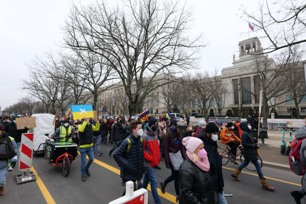 Eine große Gruppe von Menschen nimmt an einer Protestdemo auf einer Straße in Washington, D.C. teil, wobei sie Schilder und Banner schwenken und einige Fahrräder fahren, Schilder mit Stangen, Bäume und einen klaren blauen Himmel im Hintergrund.