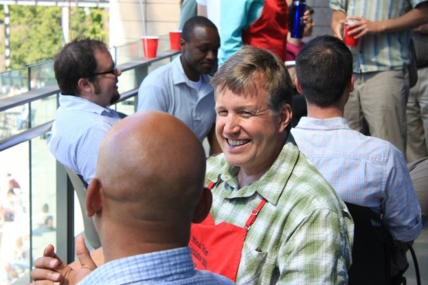 Ein Mann in einer Schürze, identifiziert als der Küchenchef der University of Texas at Austin, spricht mit einem anderen Mann auf einem Balkon, während eine Gruppe von Menschen um ihn herum sitzt und steht, mit Bäumen und einem Gebäude im Hintergrund.