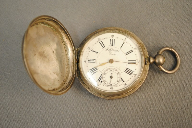 Eine silberne Taschenuhr mit einem weißen Zifferblatt mit römischen Zahlen, geschlossen mit einem Deckel, auf einer grauen Oberfläche.