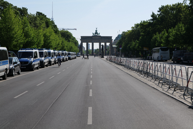 Lange Reihe von Polizeiwagen auf der Straße vor dem Brandenburger Tor in Berlin, Deutschland, mit Menschen auf Fahrrädern und in der Nähe Stehenden, Barrieren, Bäumen, einem Bogen mit Statuen im Hintergrund und dem Himmel darüber.