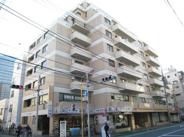 Ein mehrstöckiges Wohnhaus an einer Straßenecke, umgeben von anderen Gebäuden, Straßeninfrastruktur, Fahrzeugen, Fußgängern und Bäumen unter einem klaren blauen Himmel.