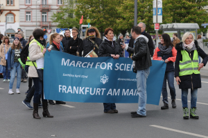 Gruppe von Menschen marschiert auf einer Straße mit einem "March for Science Frankfurt am Main"-Schild mit Bäumen, Pfählen, Schildern, Gebäuden und einem klaren blauen Himmel im Hintergrund.