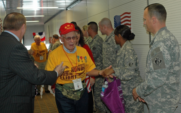 Eine Gruppe von Menschen und Soldaten steht in einem Raum mit einer Flagge an der rechten Wand und Lampen an der Decke.