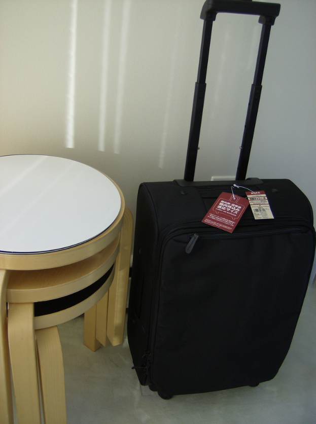 Ein schwarzer Koffer mit Etiketten oben drauf, neben einem braunen Hocker.