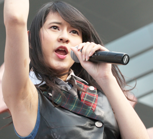 Eine Frau singt leidenschaftlich in ein Mikrofon auf einer Bühne, trägt ein weißes Hemd und einen Pferdeschwanz, hat die Augen geschlossen und ein Gebäude im Hintergrund.