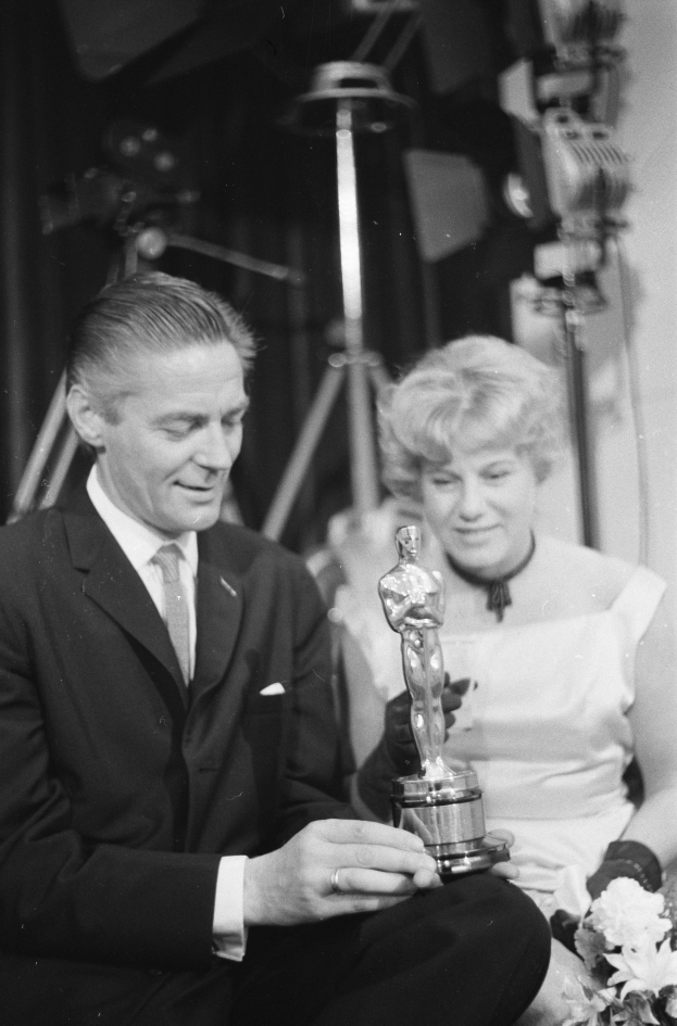 Ein Mann in einem schwarzen Blazer, weißem Hemd und Krawatte hält eine Oscar-Statue in der Hand und sitzt neben einer Frau, die einen Blumenstrauß hält, beide in eleganter Kleidung, mit Lichtern im Hintergrund, in Schwarz-Weiß.