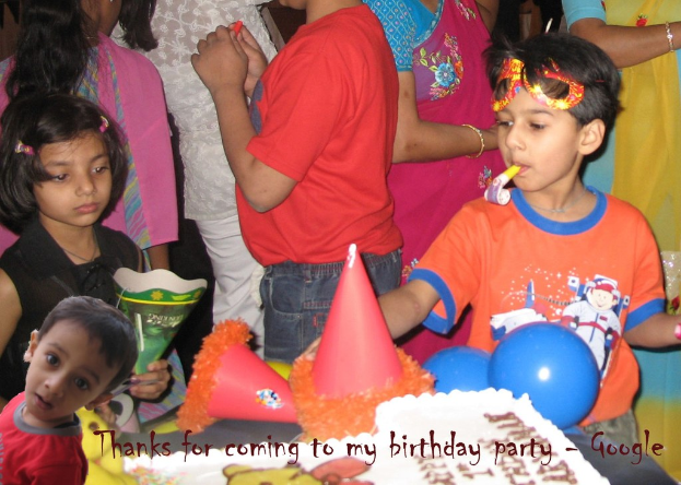 Kinder vor einem Tisch mit einem Kuchen, Luftballons und Partyhüten, mit anderen Menschen im Hintergrund.