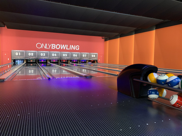 Eine Bowlingbahn mit Bowlingkugeln auf der rechten Seite, Bowlingpins in der Mitte und einer textbedeckten Wand im Hintergrund, mit der Decke oben sichtbar.
