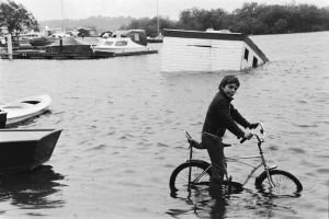 Ein junger Junge fährt mit dem Fahrrad durch eine überflutete Straße, mit Booten links, einem Haus in der Mitte und Bäumen und Himmel im Hintergrund, dargestellt in Schwarz-Weiß.