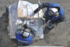 Zwei Männer in blauen Uniformen und Helmen arbeiten an einem großen Gerät, das wie eine Drohne aussieht, und halten verschiedene Gegenstände in den Händen, mit einer Plastikabdeckung auf dem Boden.