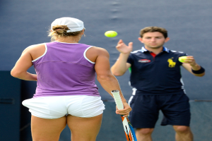 Eine Frau in einem lila T-Shirt, weißen Shorts und einer Mütze hält einen Schläger auf der linken Seite, während ein Mann in einem blauen Trikot einen Ball in ihre Richtung wirft vor einem blauen Hintergrund.