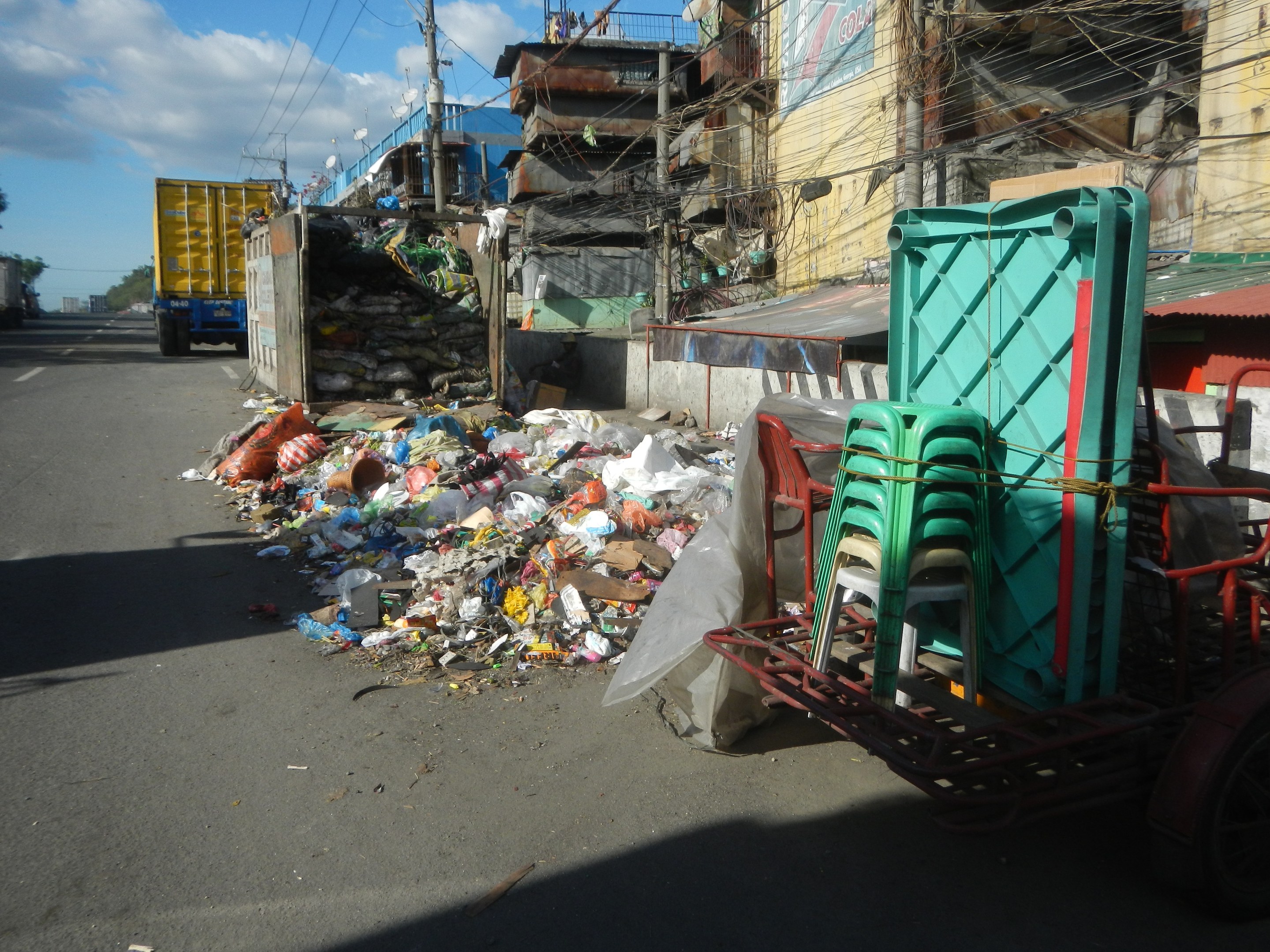 Ein Lastwagen neben einem Müllhaufen auf der Straße geparkt, mit einem Wagen rechts daneben, der mit Plastikstühlen beladen ist, und im Hintergrund Gebäude, Strommasten, Bäume und ein bewölkter Himmel.