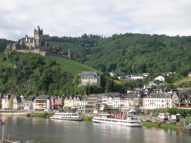 Ein malerischer Blick auf den Rhein in Deutschland mit einer Burg auf einem Hügel im Hintergrund, Booten auf dem Fluss, Fahrzeugen auf einer nahen Straße und einer bewölkten Himmel.