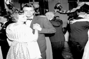 Ein Schwarz-Weiß-Foto von einem Mann und einer Frau, die auf einer Party tanzen, umgeben von lachenden Zuschauern und mit Ballons und Dekoration im Hintergrund.