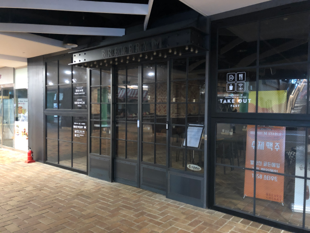 Der Eingang eines Gebäudes mit Glaswänden und -türen, ein Schild mit der Aufschrift "Brauhaus M Stable", ein Feuerhydrant auf dem Boden und Deckenleuchten, die den Bereich beleuchten.