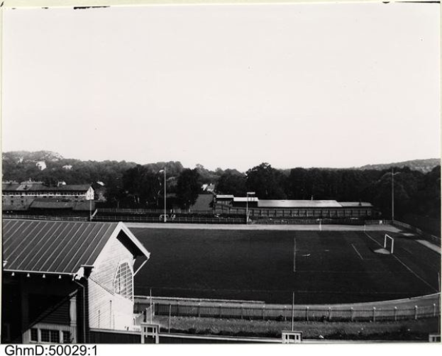 Schwarzes und weißes Foto eines Fußballfeldes mit einer Hütte links, einem umgebenden Zaun, zentralen Pfosten und Bäumen mit Himmel im Hintergrund, beschriftet mit "Gillingham Football Club, 1960er Jahre" unten.