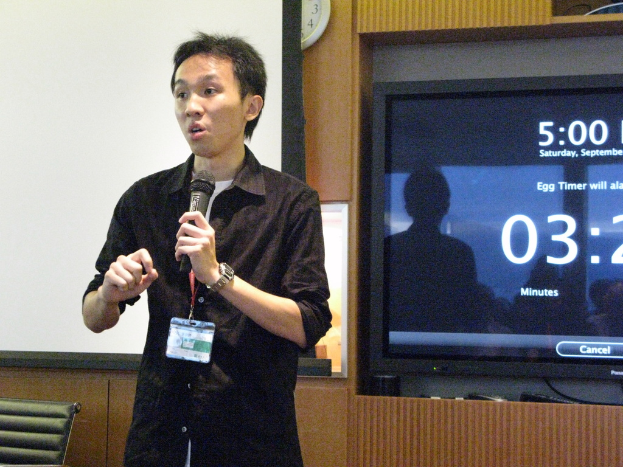 Ein Mann hält ein Mikrofon, trägt eine Tag, vor einem weißen Banner, einer Uhr an der Wand und einem Fernseher.