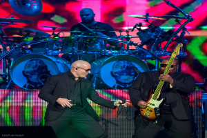 Ein Mann in der rechten Ecke spielt eine Gitarre, ein anderer Mann in einem schwarzen Blazer hält ein Mikrofon und lächelt, und ein Mann in einem schwarzen T-Shirt spielt Schlagzeug, alles vor einem bunten Hintergrund bei einem musikalischen Konzert.