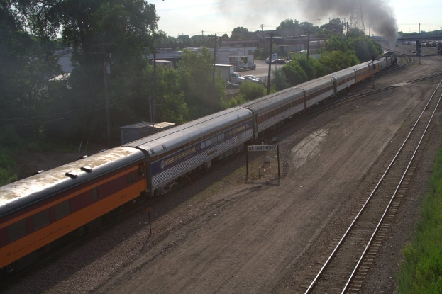Ein Zug auf Schienen mit Bäumen, Strommasten mit Kabeln, Autos und Gebäuden links und einer Brücke vorne, aus der Rauch austritt.