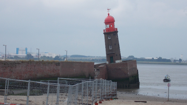 Roter Leuchtturm auf einem sandigen Strand neben Wasser mit einem Boot, eine Wand links, Bäume und Gebäude im Hintergrund unter einem klaren blauen Himmel.