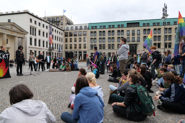 Eine Gruppe von Menschen, die auf dem Boden vor einer Menge sitzen, die Fahnen und Transparente hält, mit einer Person, die in ein Mikrofon spricht, einer Statue und Gebäuden im Hintergrund während einer Anti-Schwulen-Demo in Berlin, Deutschland.