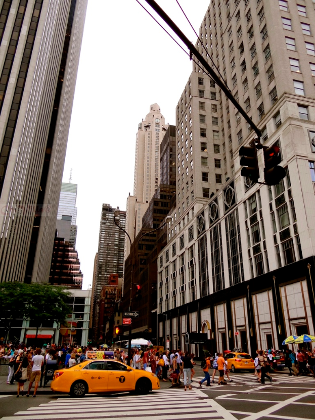 Eine Straßenszene in einer Stadt mit Gebäuden, Ampeln, Fenstern, wenigen Passanten, Autos und Himmel.