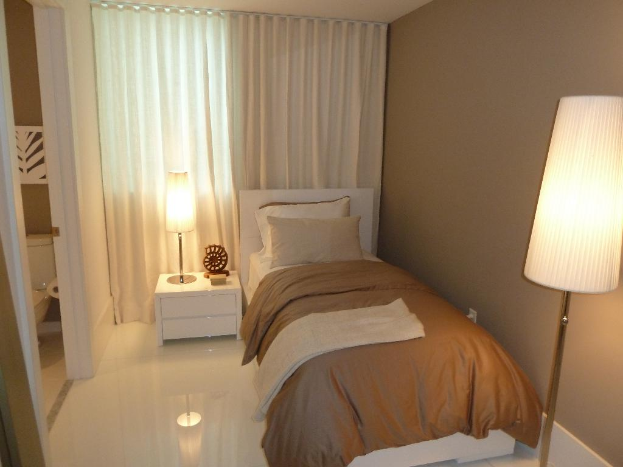 Ein Zimmer mit einem Bett, zwei Kissen, einem Nachttisch mit verschiedenen Gegenständen, einem Handtuch, einer Papierrolle, zwei Lampen, einem Vorhang und einigen an der Wand befestigten Gegenständen.