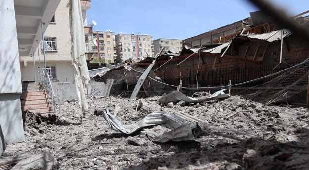 Das Bild zeigt die Auswirkungen eines israelischen Luftangriffs in Gaza-Stadt, mit einem zerstörten Gebäude in der Mitte einer Straße, umgeben von Trümmern, Metallstangen, einem Zaun, einer Treppe, benachbarten Gebäuden, Bäumen und einem bewölkten Himmel.