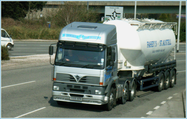 Ein Lastwagen mit Fahrer öffnet sich auf einer Straße, mit einem Van auf der Gegenfahrbahn, Bäumen und einer Brðcke im Hintergrund.