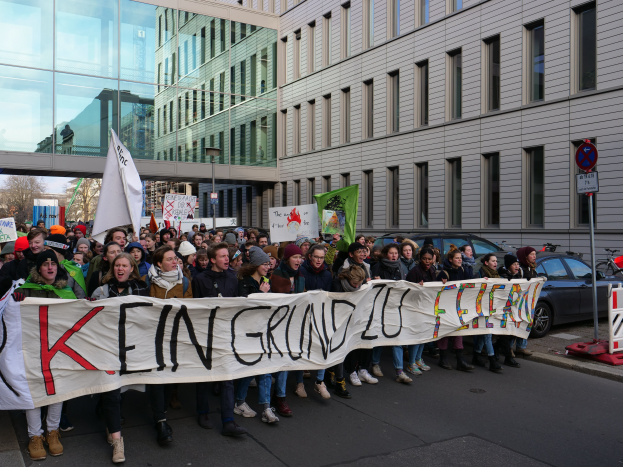 Eine Gruppe von Menschen steht auf einer Straße und hält ein Transparent mit der Aufschrift "Kein Grind Lo Feuer" in Protest, mit Gebäuden, Bäumen, Masten, Schildern, Fahrzeugen und einem klaren blauen Himmel im Hintergrund.