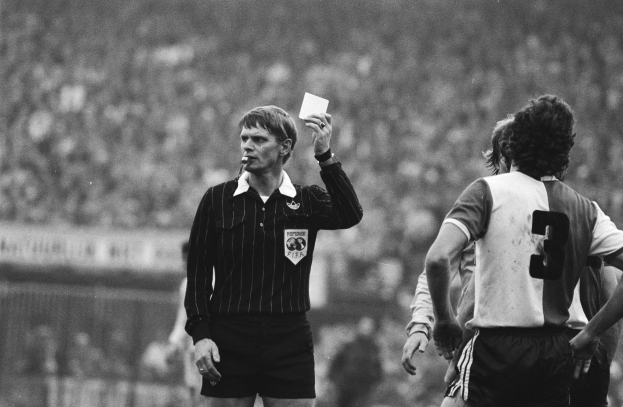 Ein Schiedsrichter in Schwarz-Weiß-Haltung hält eine Rote Karte vor einer Menge von Menschen in Fußballuniformen hoch, mit einem Zaun im Hintergrund.