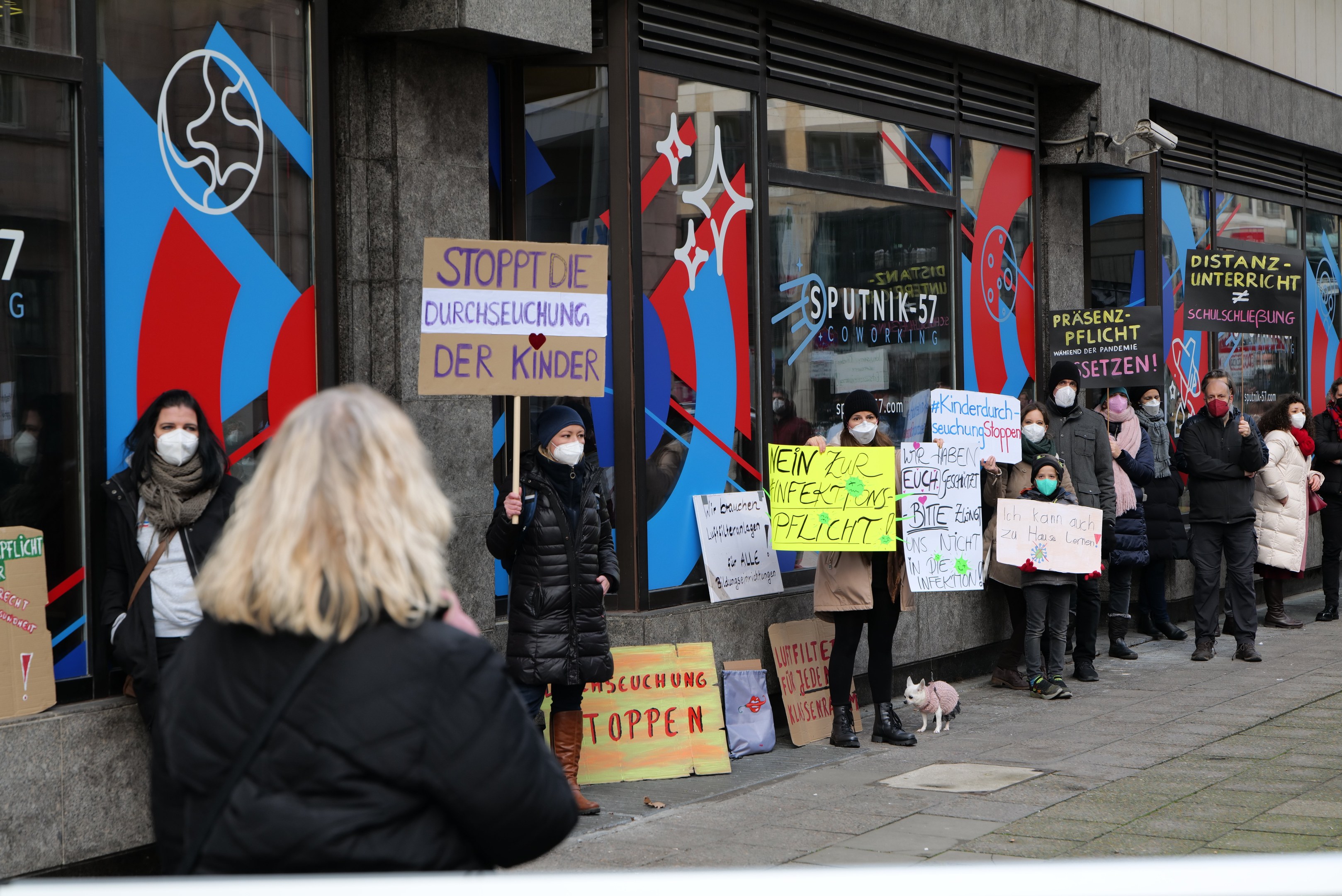 Gruppe von Menschen mit Masken und Schildern vor einem Glasfrontgebäude, mit Protesttafeln, Kameras und einem Hund unten.