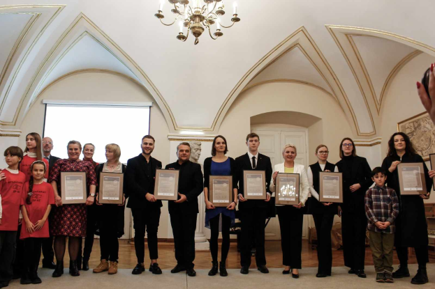 Eine Gruppe von Menschen steht nebeneinander in einem Raum, jeder hält ein Zertifikat in der Hand, mit einer Hand auf der rechten Seite, einer Wand, einer Tür, einem Bildschirm und einem Kronleuchter im Hintergrund, bei einer Preisverleihung für das akademische Jahr 2018-2019.