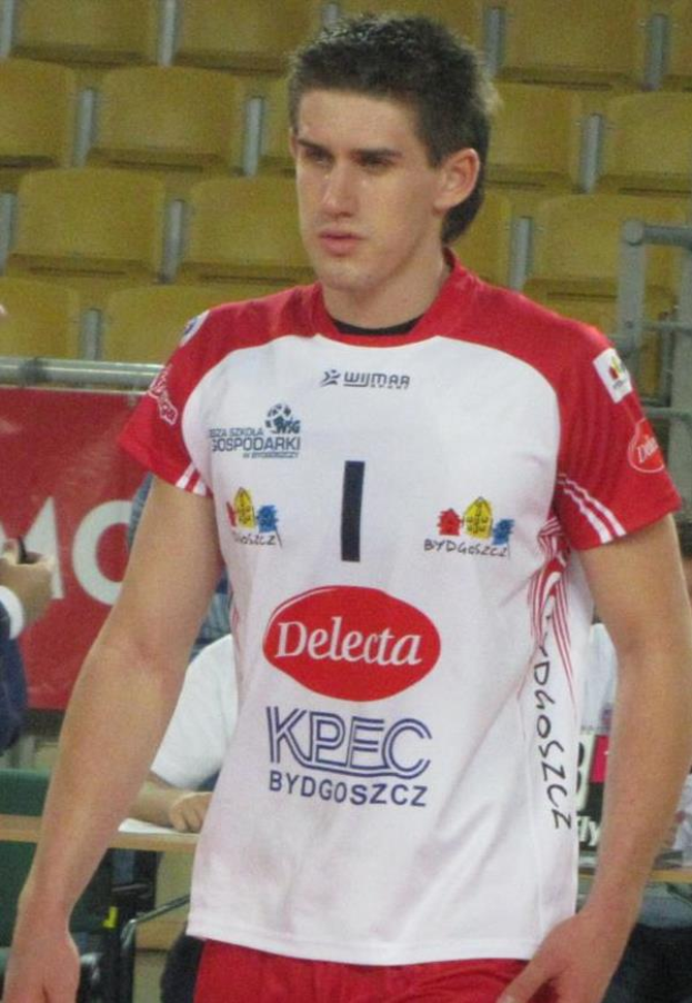 Ein Mann in einem roten und weißen Handballtrikot steht auf einem Feld, entschlossen schauend, mit Zuschauern und einem Banner mit der Aufschrift "Krasnoyarsk vs Bydgoszcz" im Hintergrund.