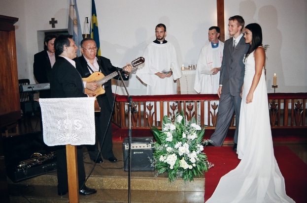 Eine HochzeitsSzene in einem Raum mit zwei Pastoren, einem Brautpaar und drei Musikern; ein Podium mit Stoff, ein Tisch mit Kerzen hinter den Pastoren, eine Kerze in einem Halter hinter dem Paar und verschiedene Gegenstände wie ein Bukett, Lautsprecher, Taschen und ein roter Teppich auf dem Boden; Flaggen, eine Wand, einen Schrank, einen Stuhl und ein Kreuz an der Wand im Hintergrund.
