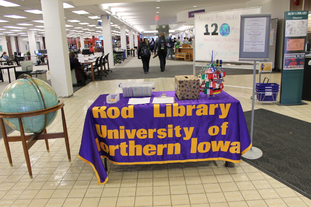 Eine Messestellung mit einem Tisch, auf dem verschiedene Landesflaggen, ein Karton, Papiere und ein Banner sowie links daneben eine Weltkugel, eine Infowand mit Menschen und rechts daneben mehrere Tische und Stühle zu sehen sind.