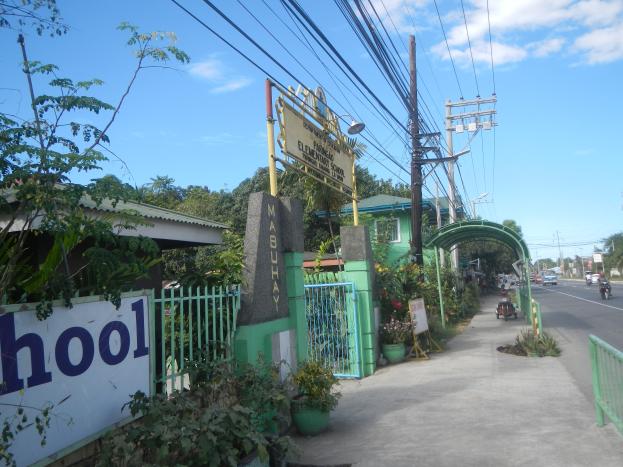 Der Eingang einer Schule mit einem Schild, auf dem 'Schule' steht, auf der Seite einer Straße, mit einem Bogen, Tor, Namensschild, Pflanzen, Bäumen, Strommasten mit Drähten und einem bewölkten Himmel im Hintergrund.