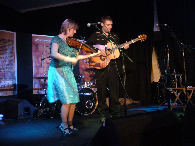 Ein Mann in schwarzem Hemd und Hose spielt Gitarre in der Mitte der Bühne, während eine Frau in einem blauen Kleid links von ihm Geige spielt, mit verschiedenen Musikinstrumenten und Lautsprechern im Hintergrund.