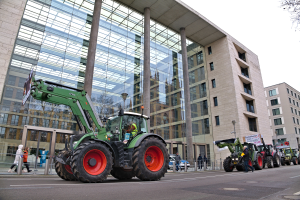 Eine Gruppe von Traktoren fährt auf einer Straße an einem Gebäude vorbei, mit Menschen auf dem Gehweg und einem Baum auf der rechten Seite, unter einem sichtbaren Himmel, wahrscheinlich an einer Demonstration teilnehmend.