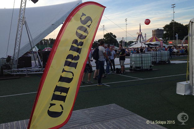 Ein buntes Karnevalsszenario mit Zelten, einem zentralen Mast, Menschen, Bäumen, Strommasten, Straßenbahnen auf der Straße und einem eisernen Stand auf der linken Seite.