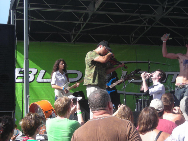 Eine Gruppe von Musikern, die verschiedene Instrumente auf einer Bühne mit einem großen schwarzen Lautsprecher links und einer grünen Fahne im Hintergrund spielen, während das Publikum davor steht und genießt.