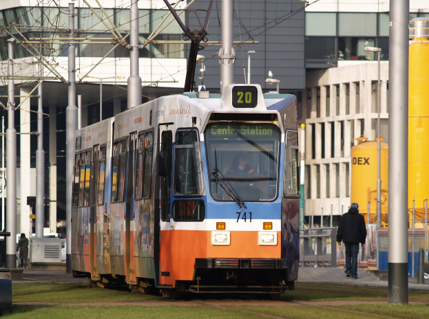 Blauer und oranger Tram auf einer Stadtstraße mit einem Passagier im Inneren, einer Person auf dem Gehweg und Gebäuden im Hintergrund.