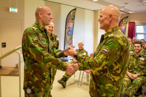 Zwei Männer in Militäruniformen geben sich die Hand vor einer sitzenden Gruppe, mit einem Tisch, Geländer, Fahnen und diversen Hintergrundelementen einschließlich einer Tür, einer Schautafel, einem Fenster mit Vorhang und Deckenleuchten, was auf eine deutsche Armeeversammlung hindeutet.