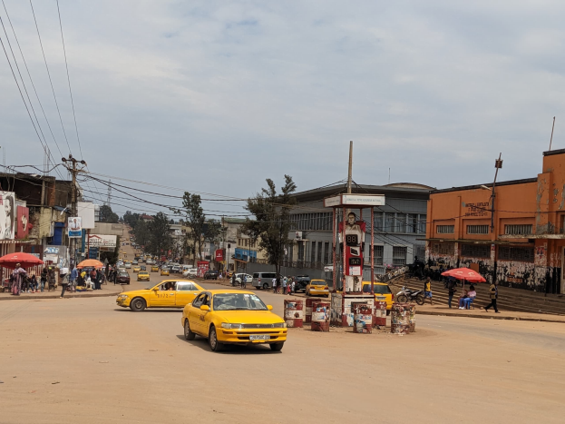 Ein gelbes Taxi fährt auf einer belebten Stadtstraße mit Gebäuden an den Seiten, während Fußgänger mit Schirmen auf dem Gehweg unter einem bewölkten Himmel gehen, mit Strommasten und Kabeln im Hintergrund.