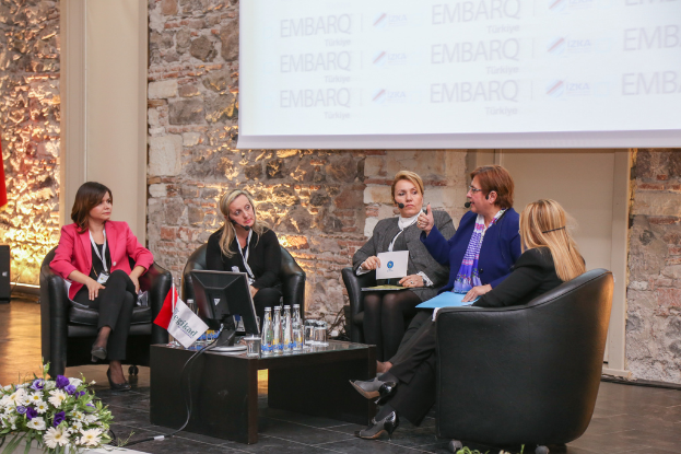 Mehrere Frauen sitzend, mit Papieren und ID-Badges, Flaschen, Gläsern, Flaggen und einer Vase auf dem Tisch sowie einem Bildschirm an der Wand.