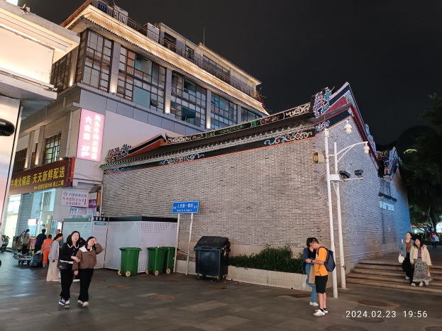 Menschen, die nachts eine Straße entlanggehen, beleuchtet von Straßenlaternen und umgeben von Gebäuden, Schildern, Mülltonnen, Pflanzen, Bäumen und einem bewölkten Himmel, einige tragen Taschen.
