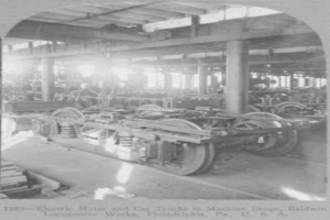 Schwarzes und weißes Foto eines Elektromotors und eines Lastwagens in einer industriellen Fabrikumgebung mit Maschinen, Säulen und einem Dach, das Text am unteren Rand enthält.