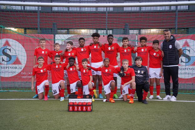 Eine Gruppe junger Männer in roten T-Shirts und schwarzen Shorts steht auf einem Fußballfeld, mit einer Kiste roter Gegenstände davor und Bannern im Hintergrund, die "FC Red Black Eglaute - Champions League" lesen.